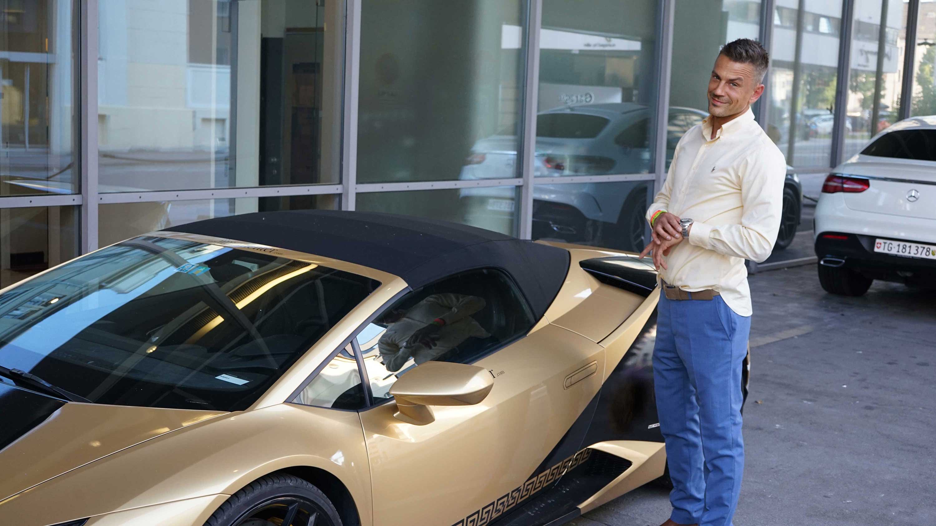 Ein lächelnder Mann in blauer Hose steht neben einem goldenen Sportwagen.